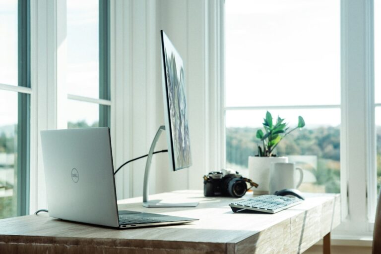 Real photograph of a modern sunlit home office workspace with laptop, external monitor, and desk accessories