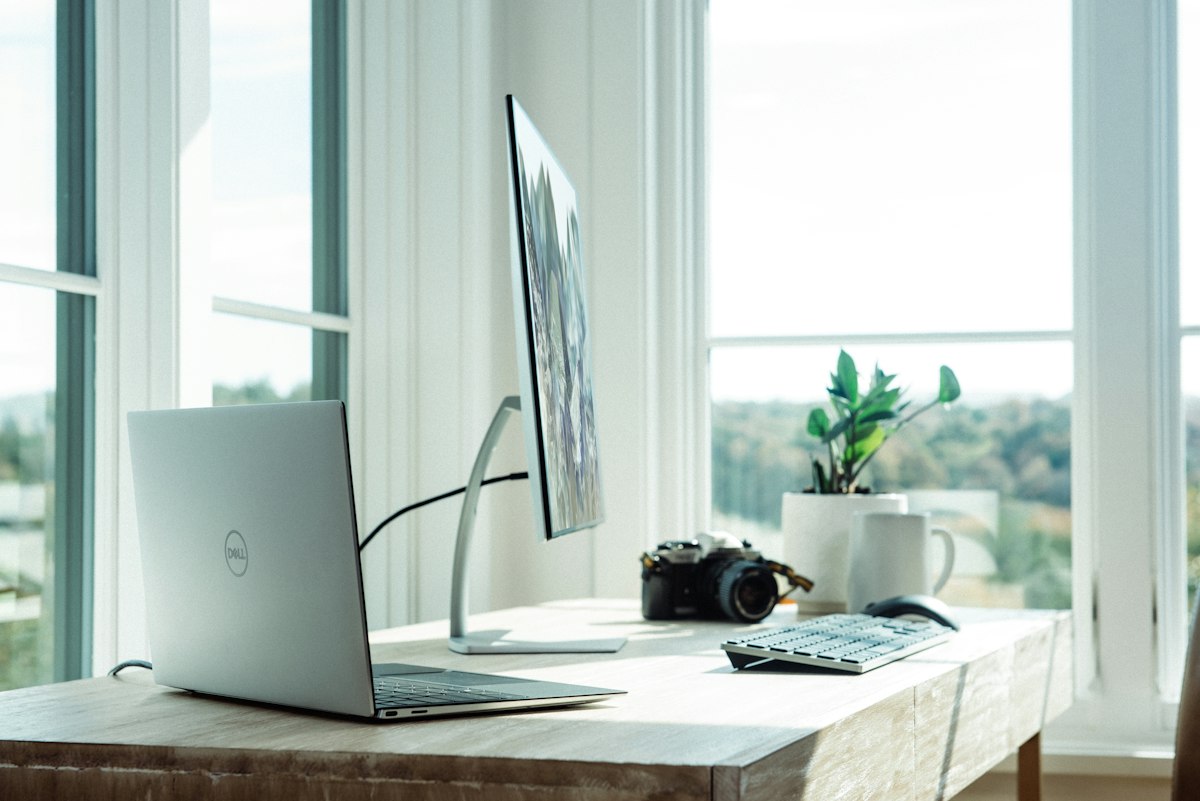 Real photograph of a modern sunlit home office workspace with laptop, external monitor, and desk accessories